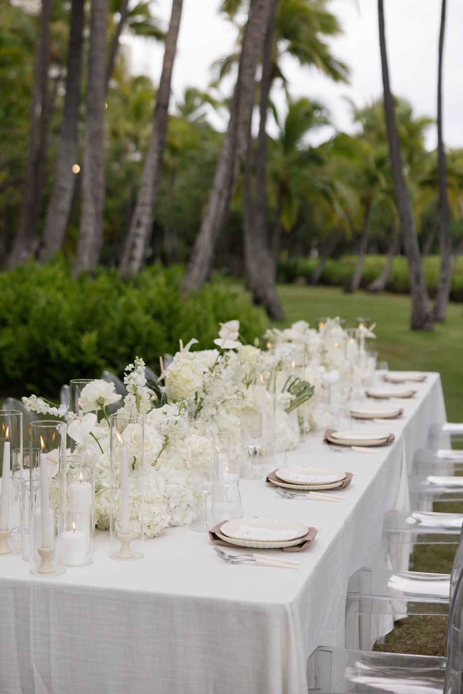 All-white floral table under coconut palms with taper candles in glass hurricanes
