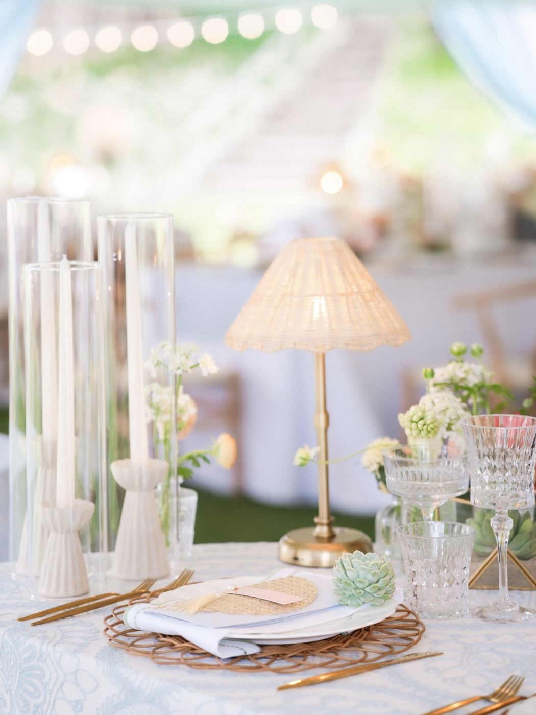 Brass rattan lamp beside an Eliza plate place setting with matte gold flatware, ceramic tapers in hurricane sleeves and crystal glassware