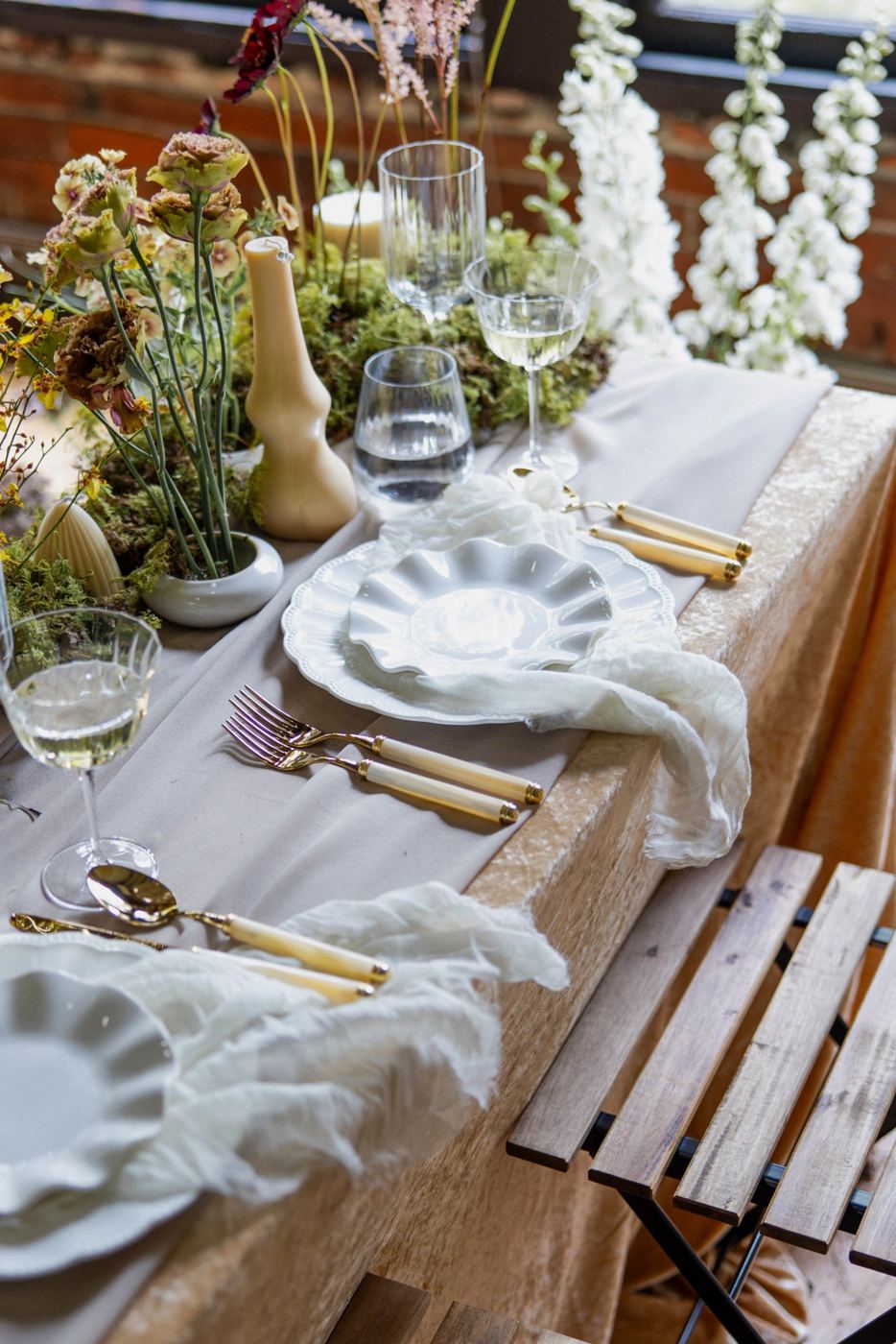 Neutral tablescape with ruffle-edge plates, pearl-handled gold flatware and ribbed glassware beside a moss and wildflower centerpiece