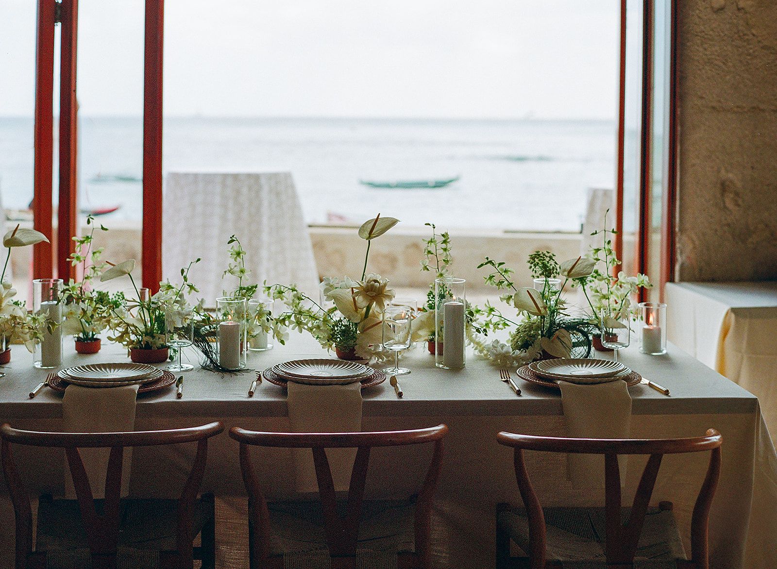 Long dinner table facing the ocean and outrigger canoes on Oʻahu, set with Zoe fluted plates on rattan chargers, gold flatware, white orchids and pillar candles