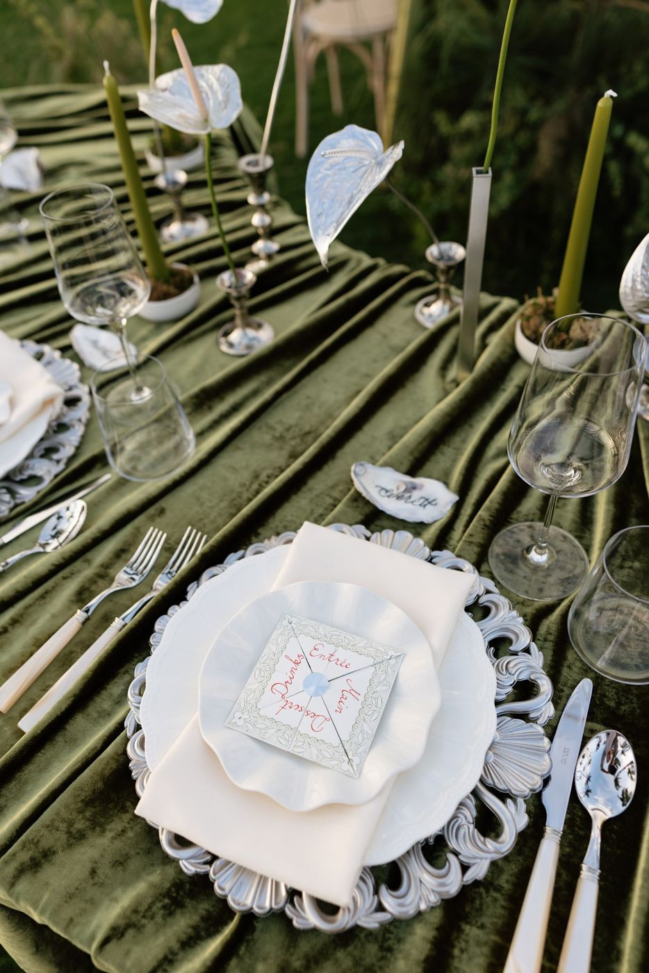 Place setting on olive velvet with an ornate silver charger, ruffle-edge plate, pearl-handled flatware, crystal glassware and silver anthuriums