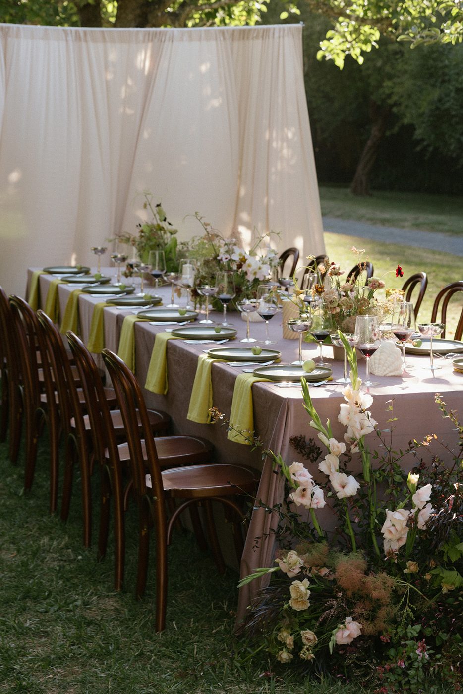 Long garden table with olive Ivy stoneware plates, chartreuse napkins, Lustra stainless coupes and wild floral centerpieces beside a draped backdrop