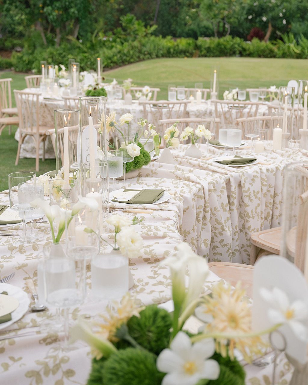 Long tables in a sage leaf-print linen with taper candles and white florals on an Oahu lawn