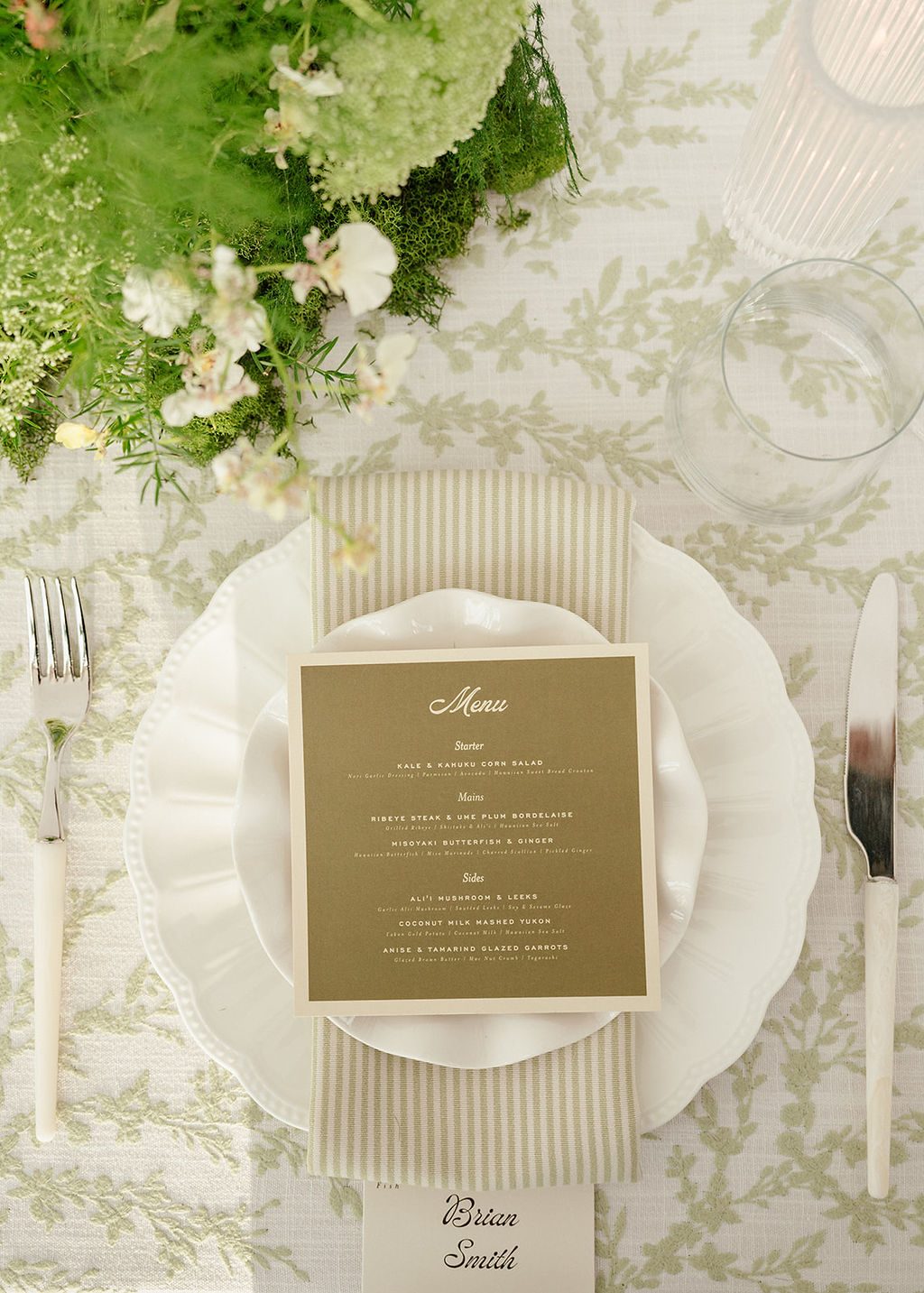 Overhead place setting with Eliza plates, an olive menu, striped napkin and Ivory polished silver flatware on a sage leaf-print linen